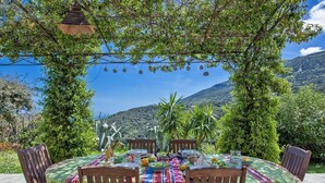 An outdoor dining area with a table set for four, surrounded by wooden chairs, under a pergola with hanging decorations. - Villa Merusellu (Sisco)