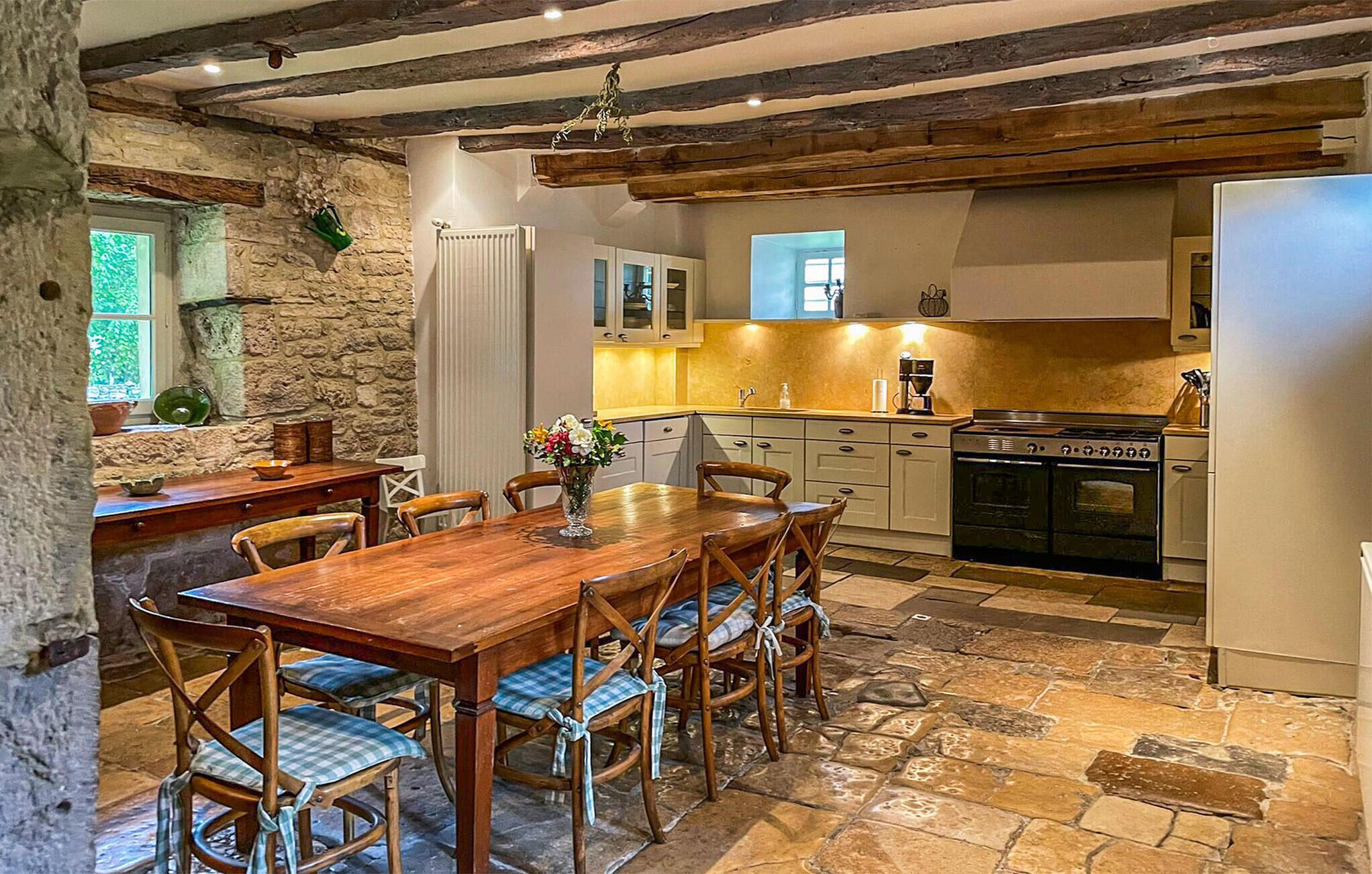A rustic kitchen with stone walls, wooden beams, a dining table with chairs, and a stove.