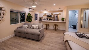 Un appartement moderne avec un canapé gris, un ventilateur de plafond, des luminaires suspendus, une cuisine équipée d’un réfrigérateur et une salle de bain visible à travers une porte ouverte.