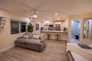 Un appartamento moderno con un divano grigio, una ventola a soffitto, lampade a sospensione, una cucina con frigorifero e un bagno visibile attraverso una porta aperta.