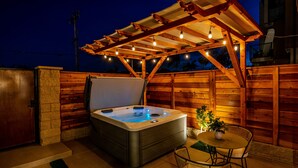 Un bain à remous sous une pergola en bois, illuminée de guirlandes lumineuses, entouré de murs en bois et agrémenté d’une table de patio avec des chaises.