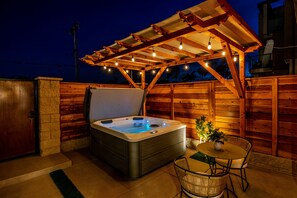Una vasca idromassaggio sotto un pergolato in legno con luci a stringa, circondata da pareti in legno e un tavolo da patio con sedie.