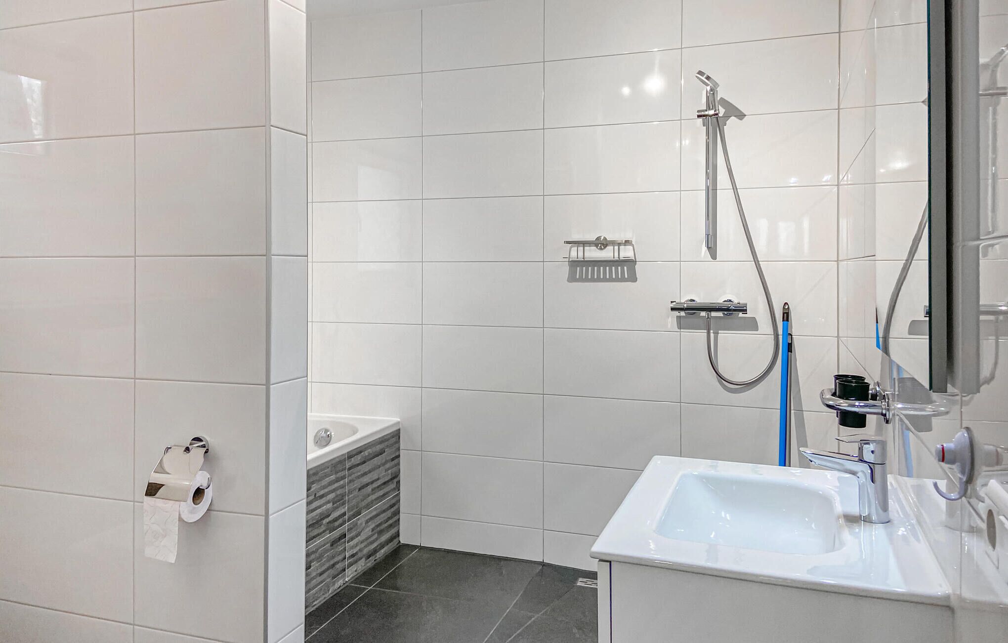 A modern bathroom with white tiles, a sink, a shower with a handheld showerhead, and a toilet paper holder.