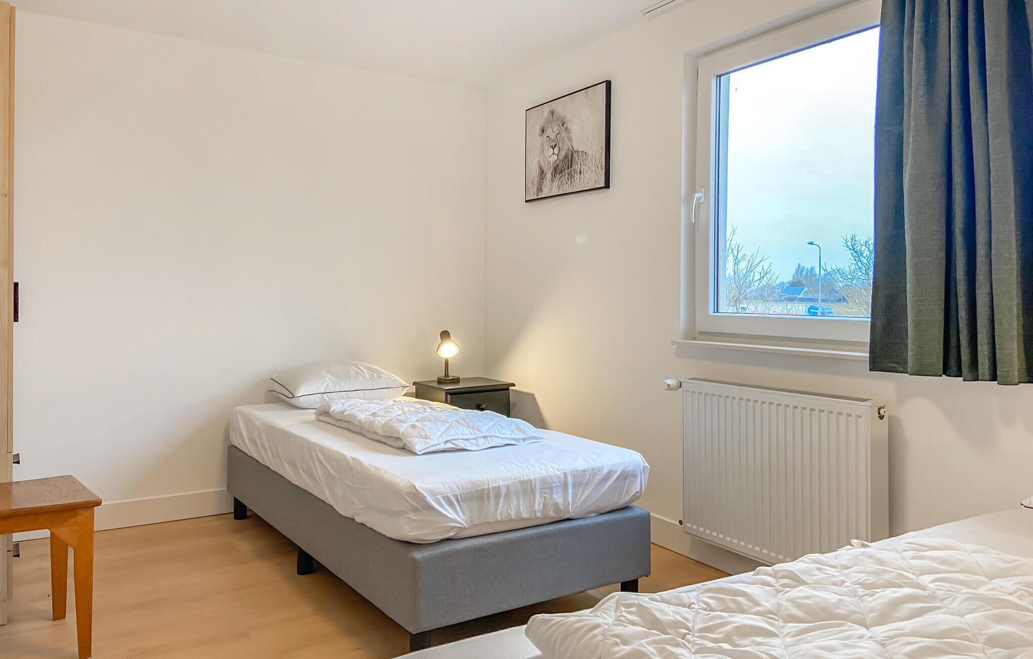 A bedroom with a bed, bedside table, lamp, window, radiator, and a view of the outside.