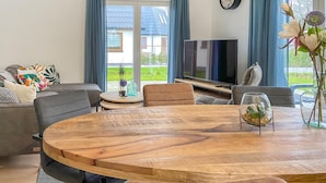 Una sala de estar moderna con una mesa redonda de madera, sofás grises, un televisor de pantalla plana y un gran ventanal con cortinas azules.