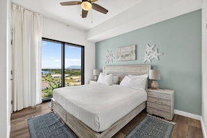 A bedroom with a large bed, two nightstands, a ceiling fan, and a view of a cityscape through a sliding door. - Origin 803: Gulf-View 1BR w/ Sunset Corner Balcony! (Panama City Beach)