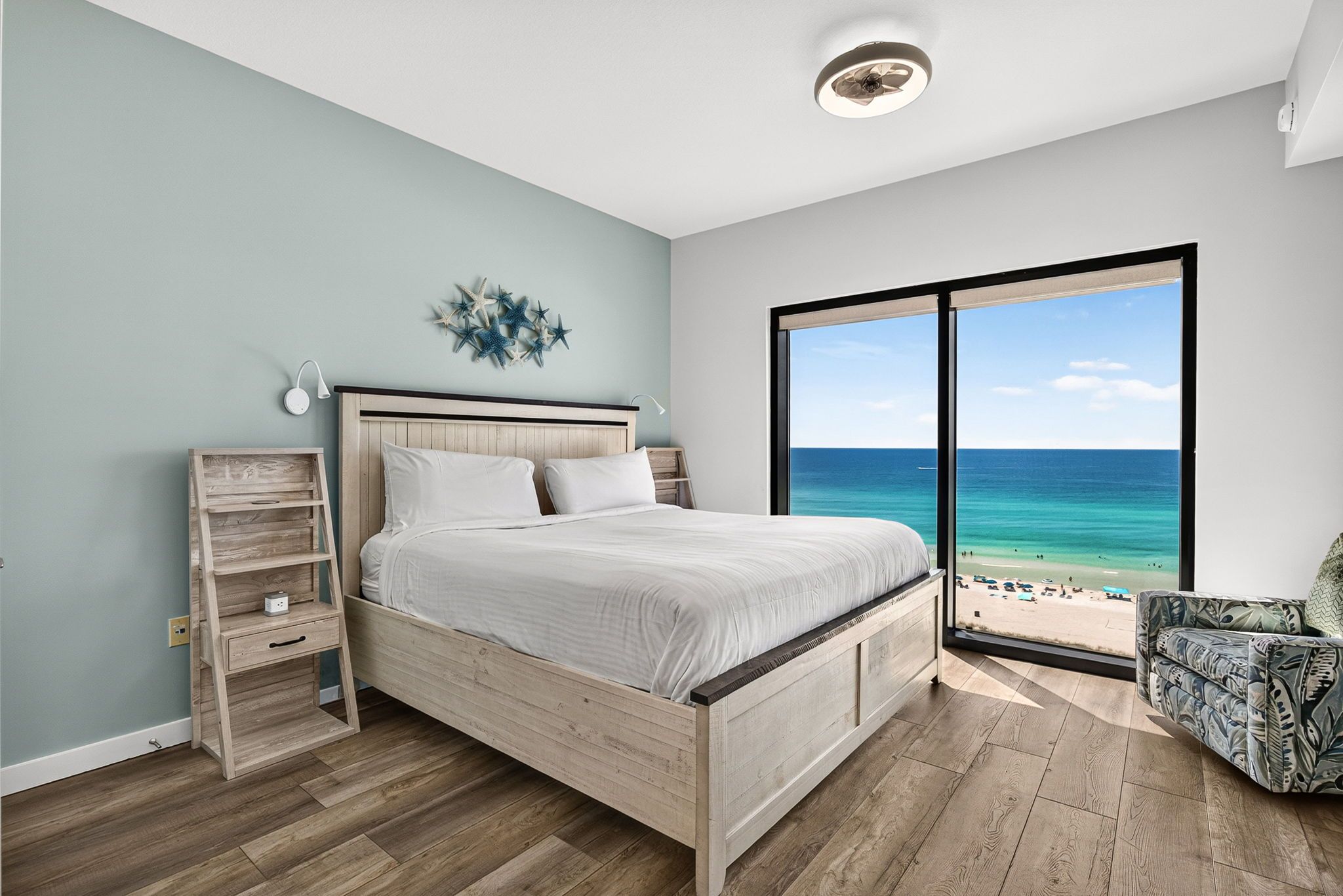 A bedroom with a bed, a nightstand, a wall-mounted lamp, a starfish decoration, and a large window with a beach view.