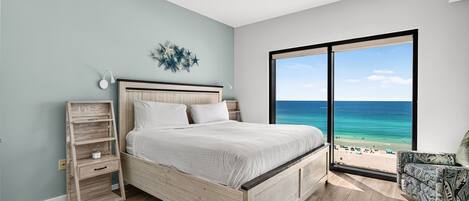 A bedroom with a bed, a nightstand, a wall-mounted lamp, a starfish decoration, and a large window with a beach view.