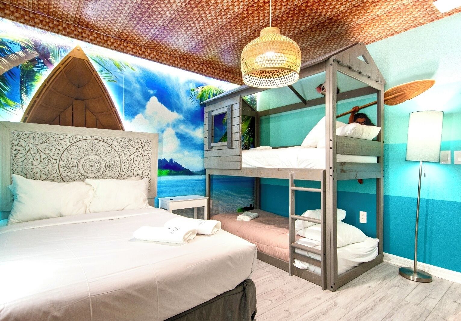 A room with a bed, bunk beds, a ceiling fan, and a mural of a beach scene.
