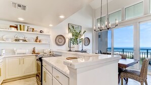 A modern kitchen with white cabinetry, a marble island, and a dining area with a view of the ocean.
