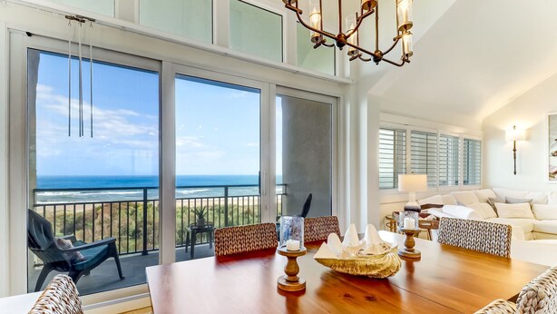 A spacious living room with a large wooden table, wicker furniture, and a view of the ocean through large windows.