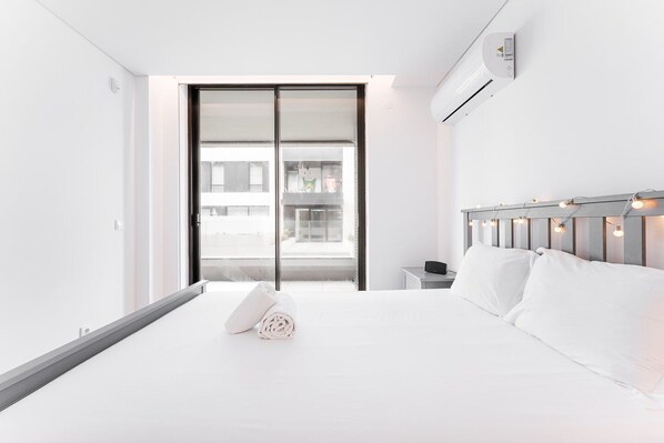 A modern hotel room with a large bed, a nightstand, a wall-mounted air conditioner, and a sliding glass door leading to a balcony.