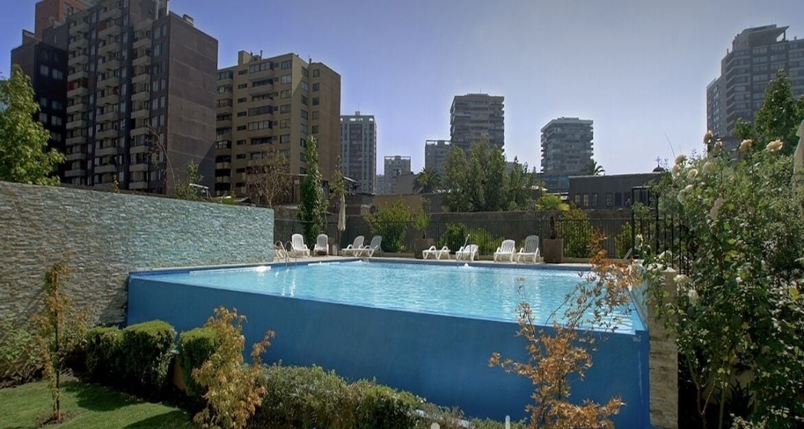A swimming pool with lounge chairs, surrounded by greenery and tall buildings in the background.