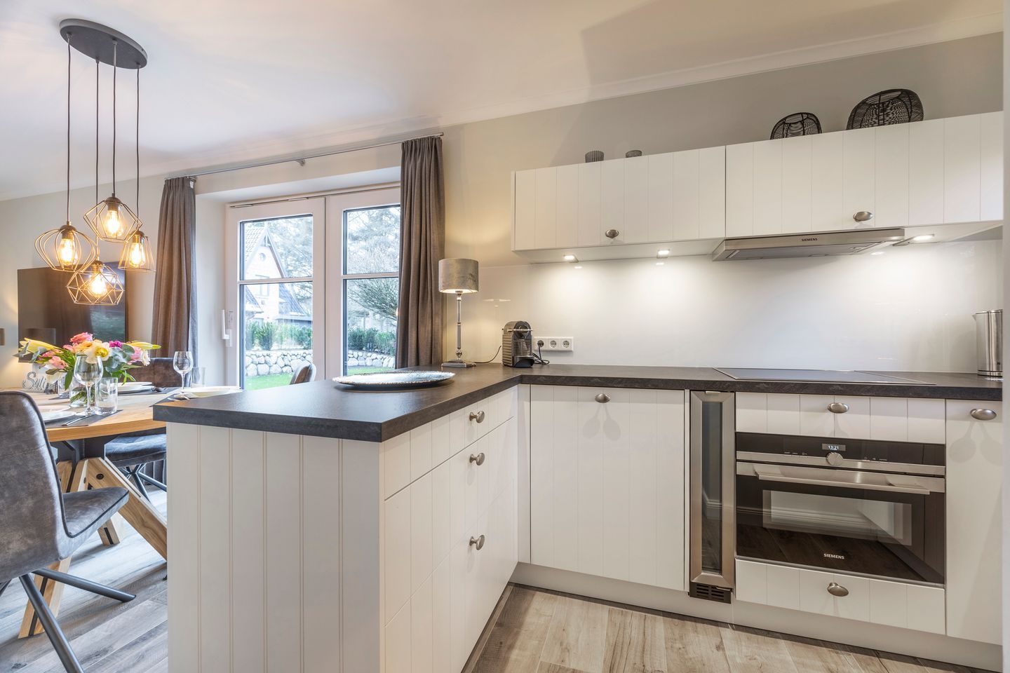 A modern kitchen with white cabinetry, a built-in oven, and a dining area with a table and chairs.