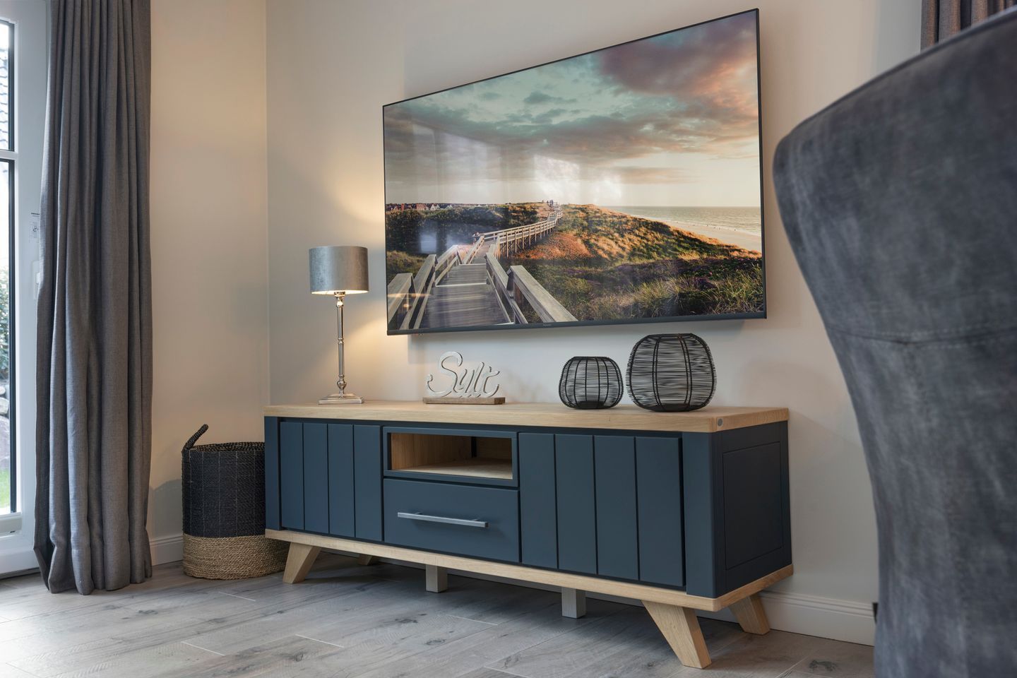 A modern living room with a large TV mounted on the wall, a dark blue cabinet with drawers, a lamp, and decorative items.