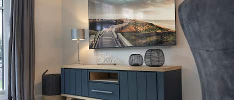 Una sala de estar moderna con un televisor grande montado en la pared, un mueble azul oscuro con cajones, una lámpara y elementos decorativos.