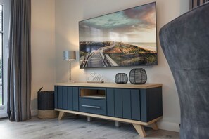 Ein modernes Wohnzimmer mit großem, an der Wand befestigten Fernseher, einem dunkelblauen Schrank mit Schubladen, einer Lampe und dekorativen Accessoires.