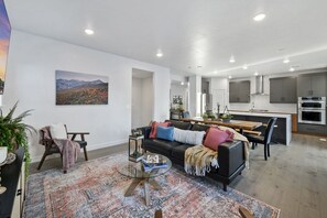 A modern living room with a black leather sofa, a glass coffee table, and a patterned rug. There is a dining area with a wooden table and chairs, and a kitchen with stainless steel appliances. - Park City Ski/Bike House (Summit County)