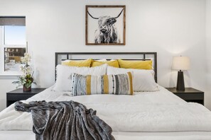 A neatly made bed with white and yellow pillows, a framed artwork above it, and a bedside lamp on a nightstand. - Park City Ski/Bike House (Summit County)