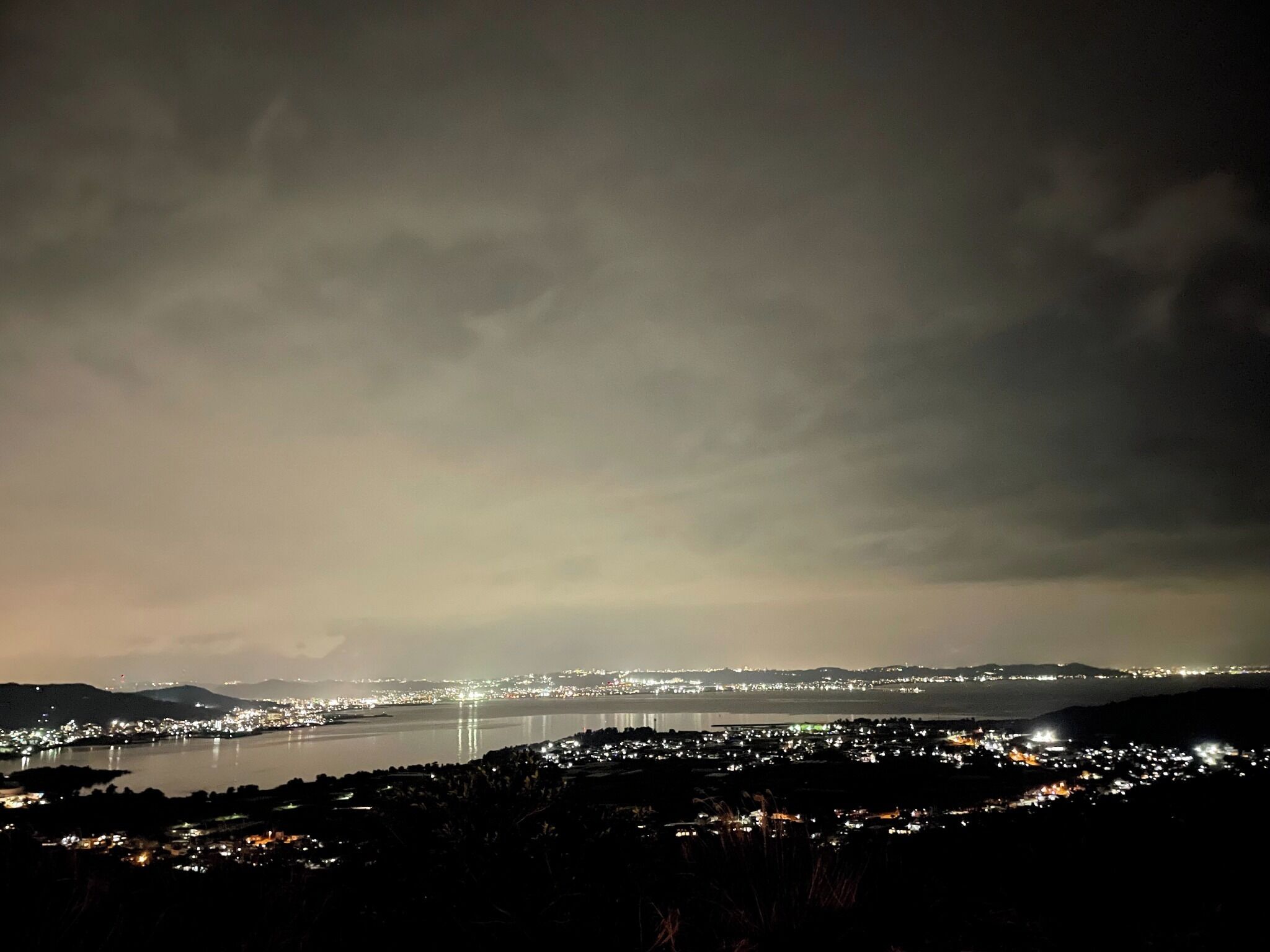 Vista nocturna de una ciudad costera con una extensa masa de agua, luces urbanas y montañas a lo lejos.