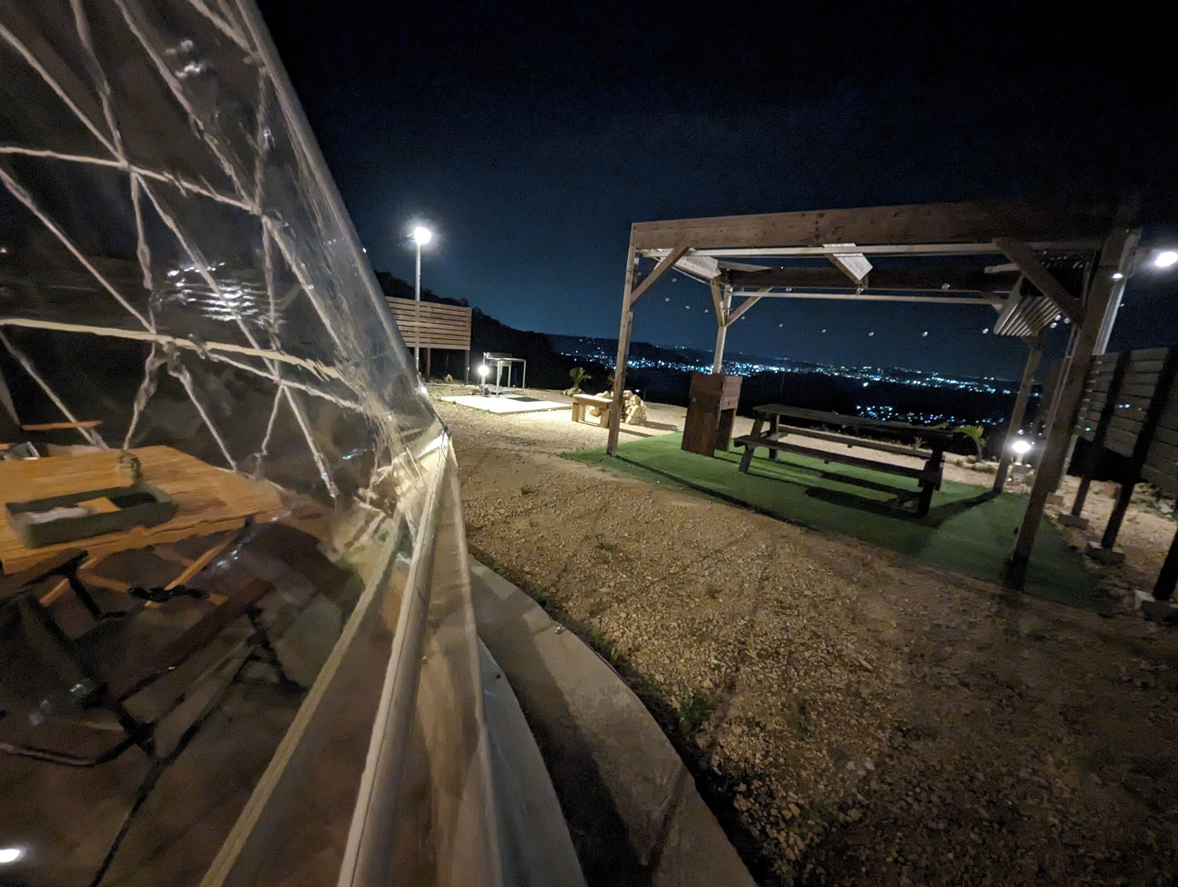 A covered outdoor area with a wooden table and benches, a pergola, and a view of the city lights in the distance.