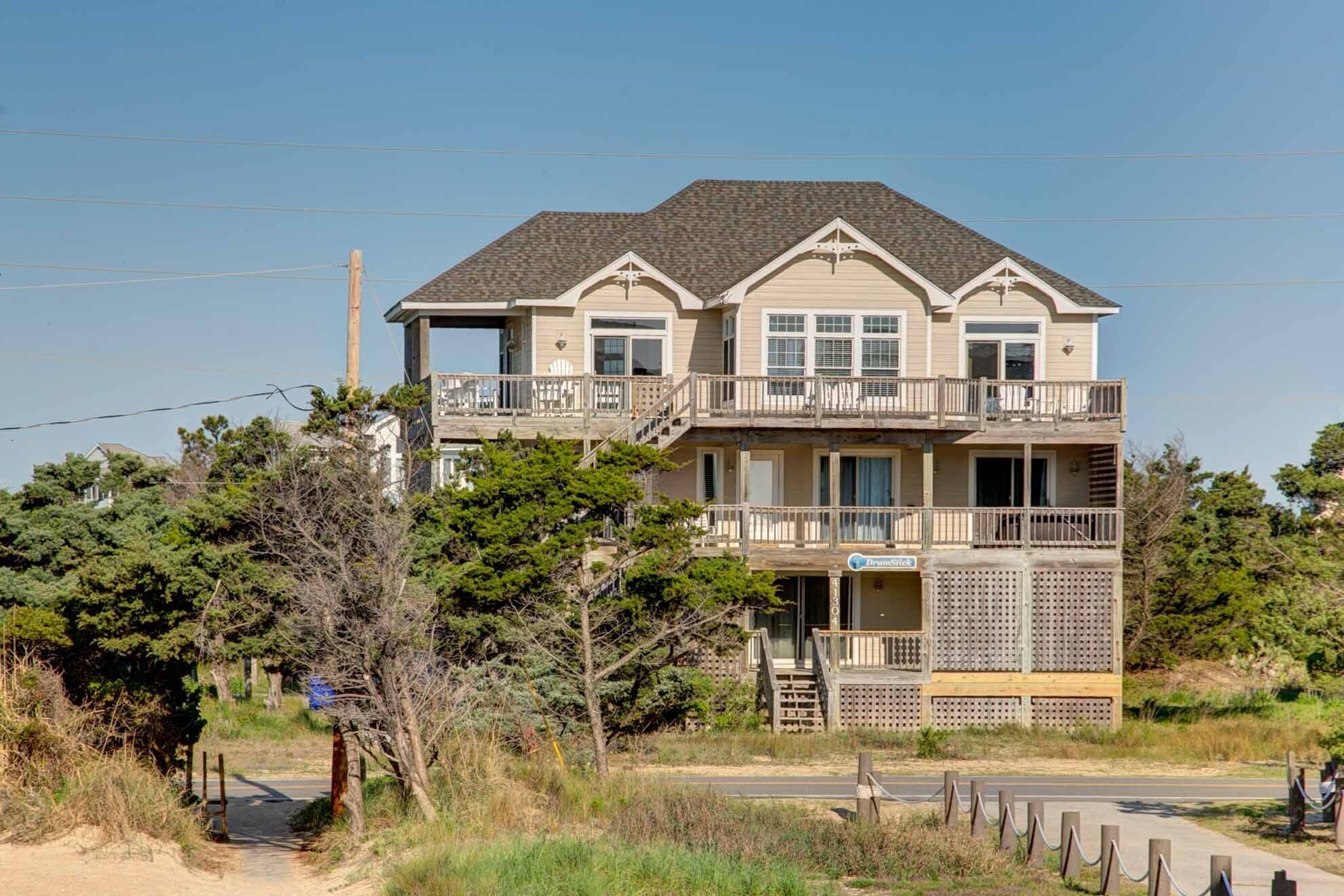 A two-story beachfront house with a balcony on the upper floor, a wooden deck, and a sign indicating 'Drumstick'.