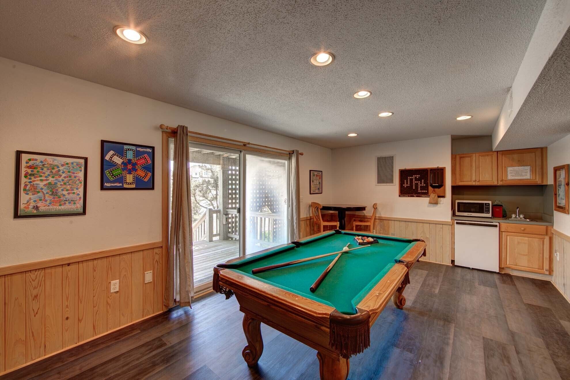 A pool table in a room with wooden paneling, a kitchen area with wooden cabinets, and a dining table with chairs.