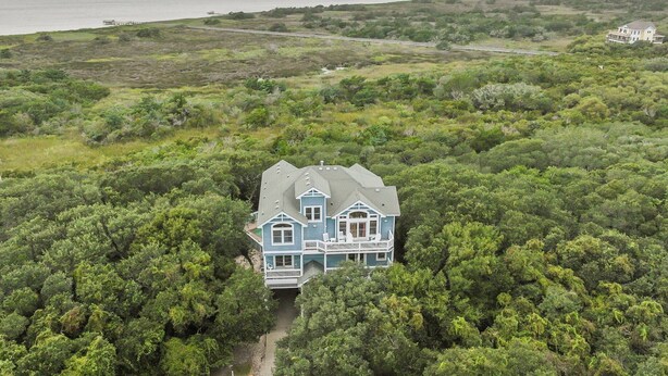 A large, two-story house with a prominent porch and multiple windows, surrounded by dense greenery and a view of the ocean. - 7725- Sun Kissed - Avon Retreat | Pool, Hot Tub & Theater Room (Avon)