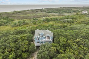 A large, two-story house with a prominent porch and multiple windows, surrounded by dense greenery and a view of the ocean. - 7725- Sun Kissed - Avon Retreat | Pool, Hot Tub & Theater Room (Avon)