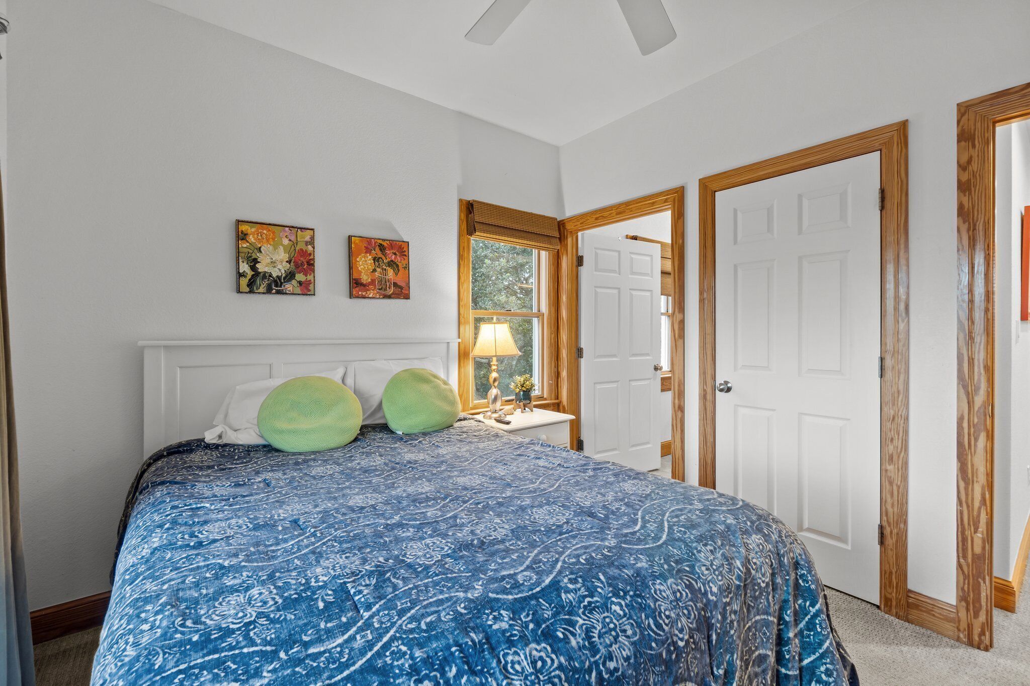 A bedroom with a large bed, two wall-mounted paintings, a window with blinds, a nightstand with a lamp, and two doors.