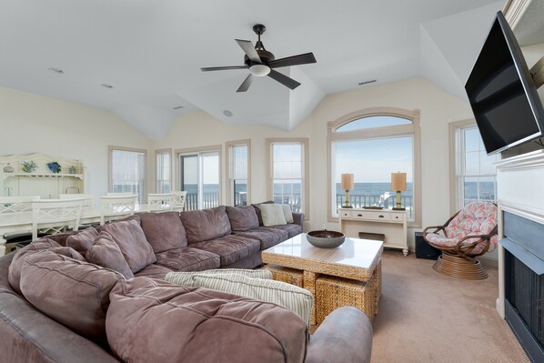 A spacious living room with a large sectional sofa, a dining area with white furniture, and a view of the ocean through large windows.