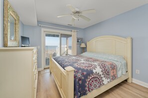 A bedroom with a bed, a ceiling fan, a dresser, a TV, and a view of the outdoors.