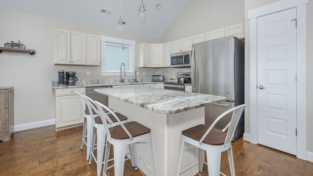 A modern kitchen with white cabinetry, a large island with bar stools, stainless steel appliances, and a window above the sink. - 5176- Sandy Bottoms - Coastal Escape | Dog Friendly + Hot Tub (Kill Devil Hills)