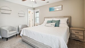 A bedroom with a bed, two chairs, a nightstand, a ceiling fan, and a window with blinds. - 5176- Sandy Bottoms - Coastal Escape | Dog Friendly + Hot Tub (Kill Devil Hills)