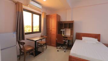 Habitación de hotel pequeña con cama, escritorio con silla, armario y ventana con cortinas.