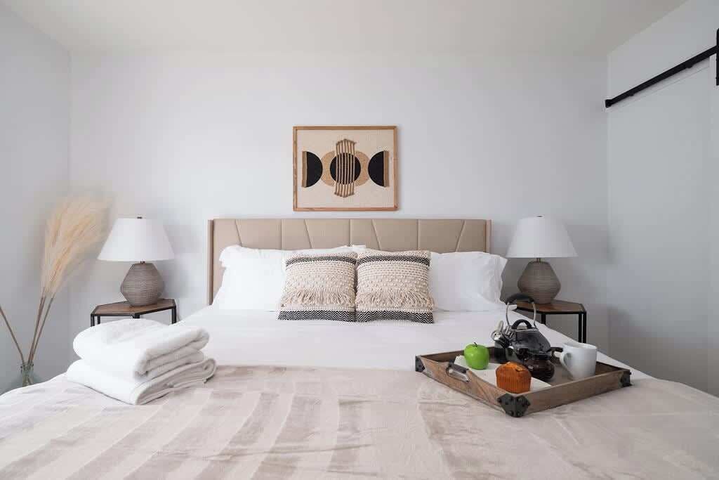 A neatly made bed with a wooden headboard, two bedside lamps, a tray with a mug, a green apple, and a cup.