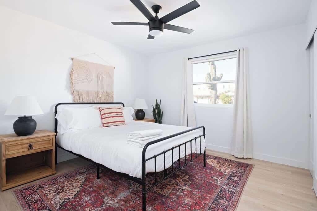 A bedroom with a bed, a ceiling fan, a window with curtains, a nightstand with a lamp, and a patterned rug.