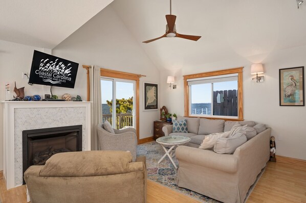 A cozy living room with a fireplace, a sofa, a chair, and a television displaying 'Coastal Carolina Vacations and Sales'. - OBX soundfront, private pier, 5 min to beach, easy to kayak & paddleboard (Nags Head)
