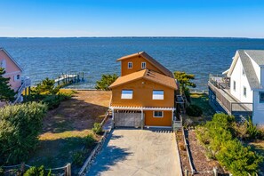 A two-story house with a garage, a wooden deck, and a view of the water. - OBX soundfront, private pier, 5 min to beach, easy to kayak & paddleboard (Nags Head)