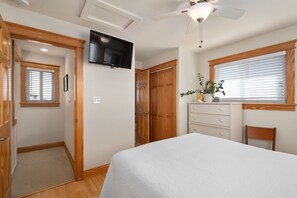 Une chambre avec un lit, un ventilateur de plafond, un téléviseur fixé au mur, des portes en bois, une fenêtre avec des stores et une petite plante sur le rebord de la fenêtre.