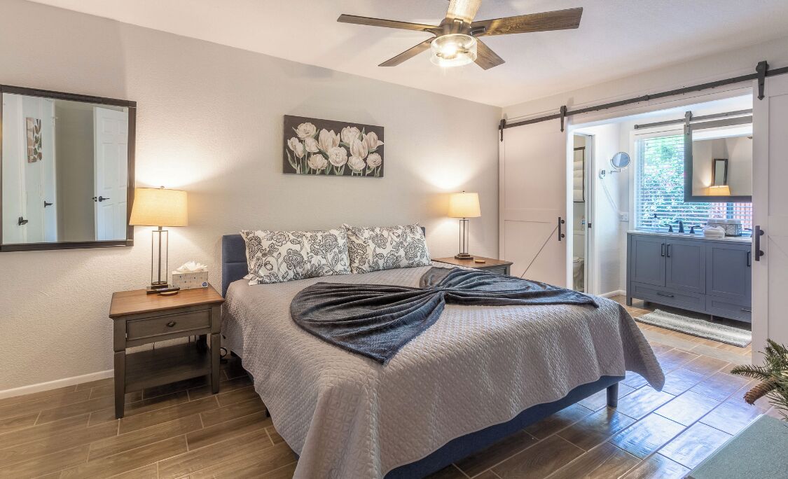 A bedroom with a bed, two bedside lamps, a ceiling fan, a large mirror, and a door leading to a bathroom.