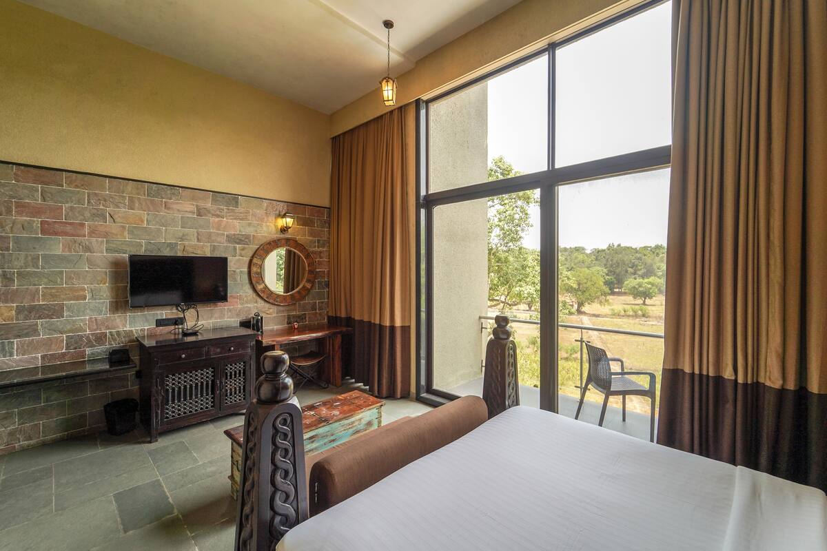 A hotel room with a large window, a flat-screen TV mounted on a stone wall, a wooden bed, and a small desk with a chair.