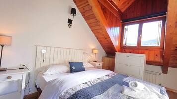 Een slaapkamer met een schuine plafond, een bed met een blauw kussen, een nachtkastje met een lamp, een dressoir en een raam met uitzicht.