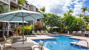 Un área de piscina con sillones de descanso y sombrillas, rodeada de exuberante vegetación y un edificio de varios pisos.