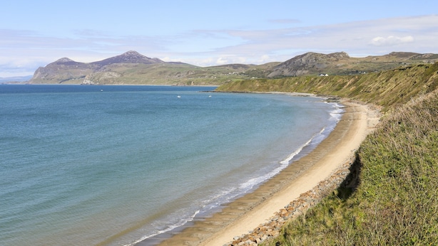 Un paysage côtier avec une plage de sable, une eau bleue et limpide, et des collines douces en arrière-plan.