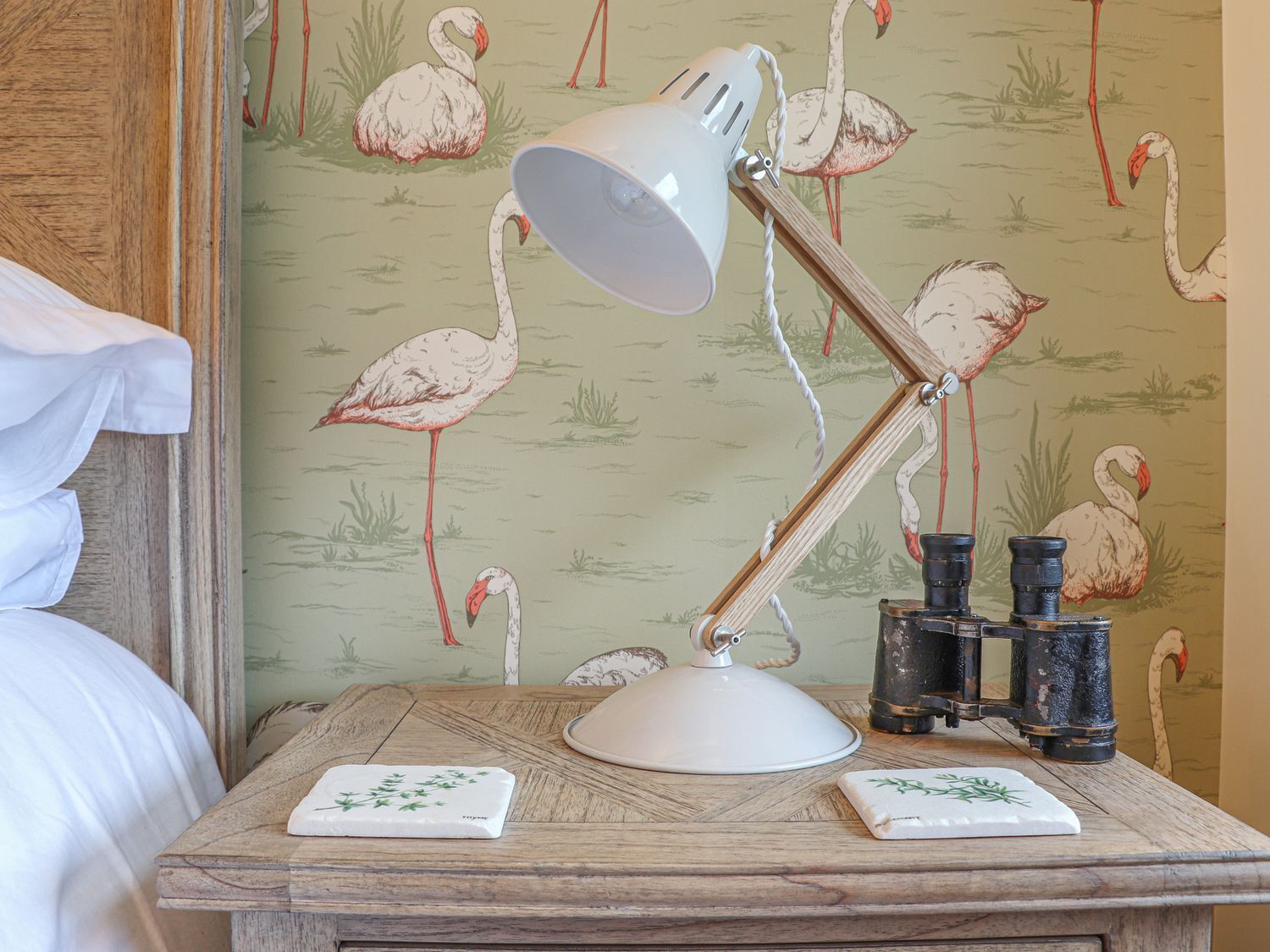 A wooden bedside table with a white lamp, two glasses, and a white plate with a green design. The wall has a wallpaper pattern of flamingos.