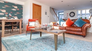 A cozy living room with a wooden coffee table, a leather sofa with colorful cushions, a dining area with a wooden table and chairs, and a fish-patterned wall. - The Old Smokehouse (St Austell)