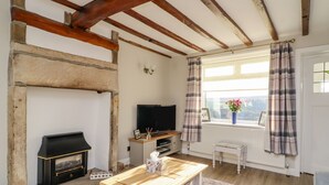 A cozy living room with a fireplace, a television, a wooden coffee table, and a view of the outdoors through a window with curtains. - Sarah's Cottage (Keighley)