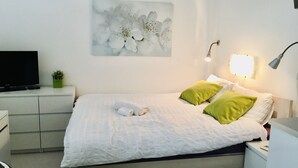 A neatly made bed with white and green pillows, a flat-screen TV on a white dresser, a small green plant, and a wall-mounted light fixture. - Hotel Flat in the Heart of Prague (Prague)