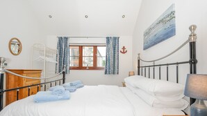 Uma cama bem arrumada com lençóis brancos, uma mesa de cabeceira de madeira com um abajur e vista para o mar através de uma janela com cortinas azuis.